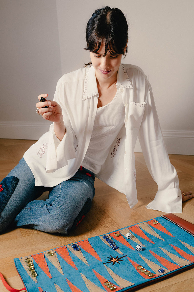 A woman in a white blouse and blue jeans kneels on a wooden floor, holding a small object over a colorful backgammon board with orange, blue, and red triangular points and playing pieces.